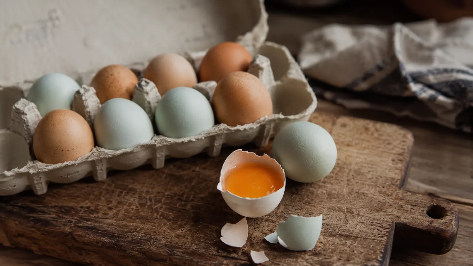 A carton of freshly collected backyard eggs with deep orange yolks visible through a cracked shell on a wooden board
